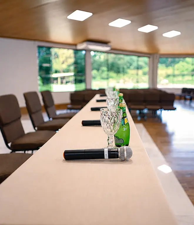 Mesa diretora do auditório Tuiuiú com microfones e flores, e a parede de vidro ao fundo